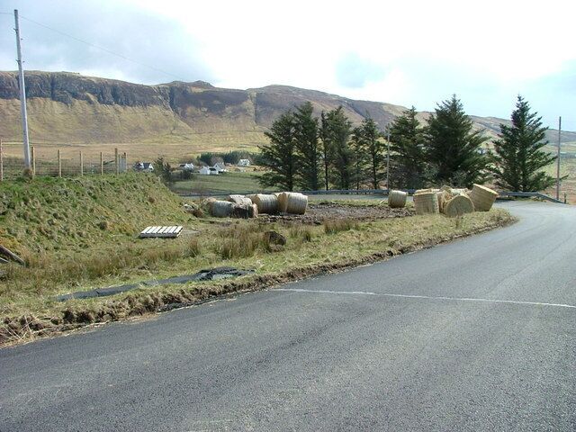 A rural road with hay bales, a fence, and a mountainous landscape in the background.