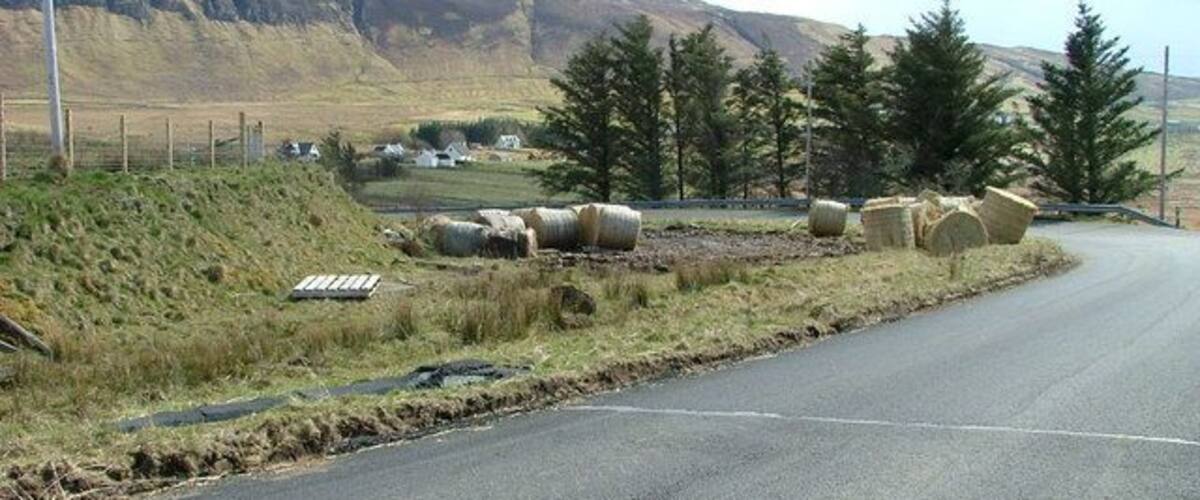 A rural road with hay bales, a fence, and a mountainous landscape in the background.