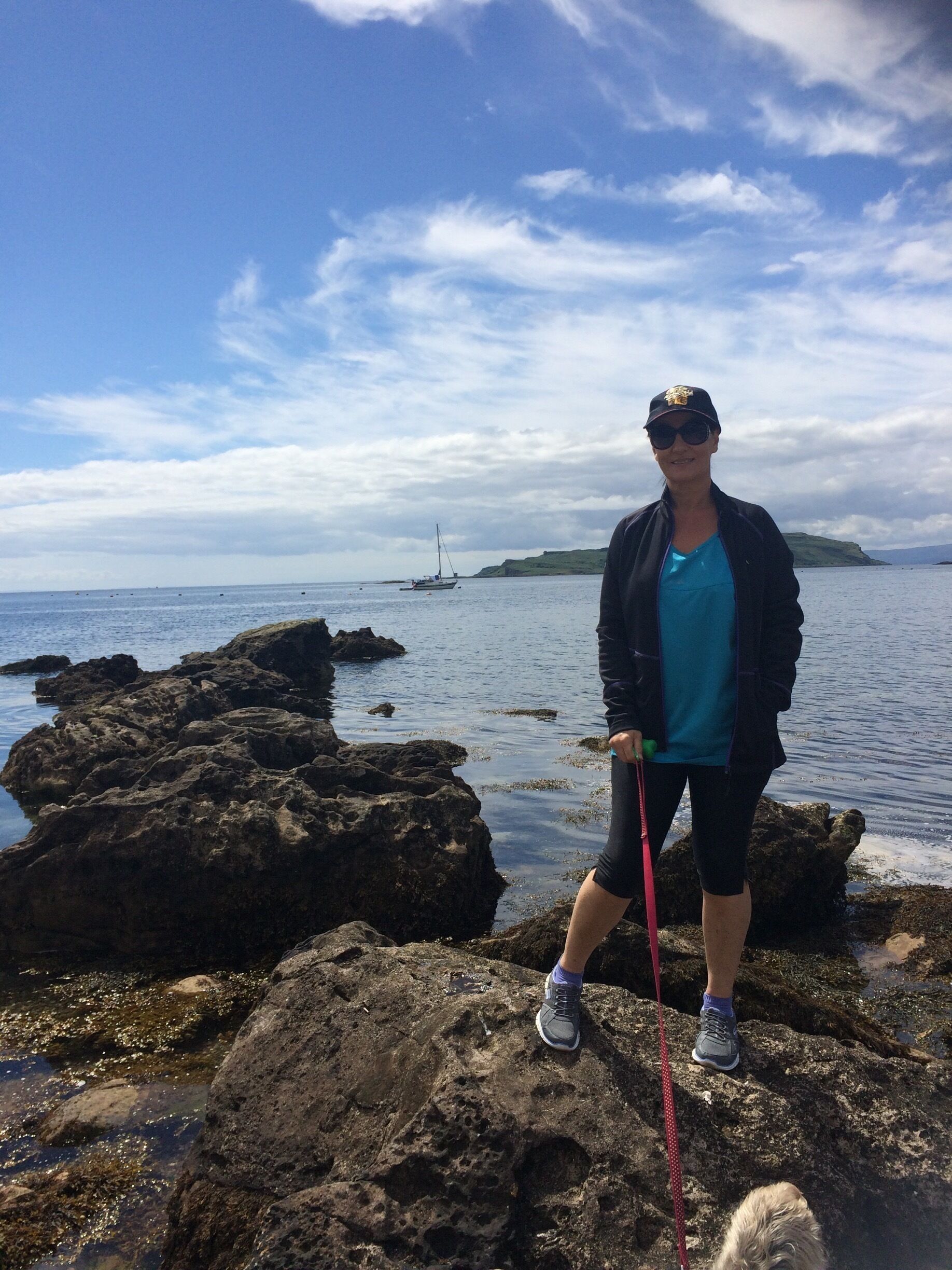 A person standing on rocky terrain near a body of water with a sailboat in the distance.