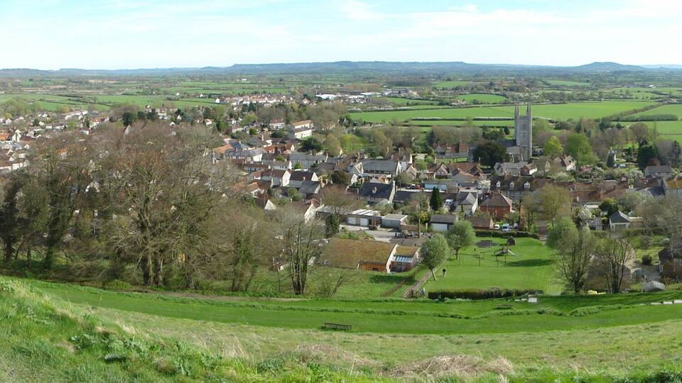 A village nestled in a valley with a prominent church tower, surrounded by green fields and hills.