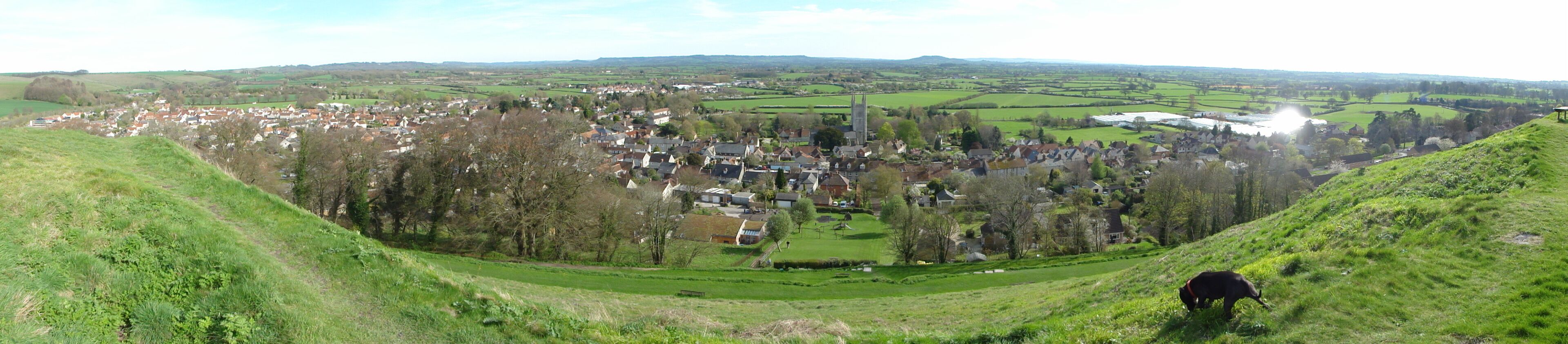 A village nestled in a valley with a prominent church tower, surrounded by green fields and hills.