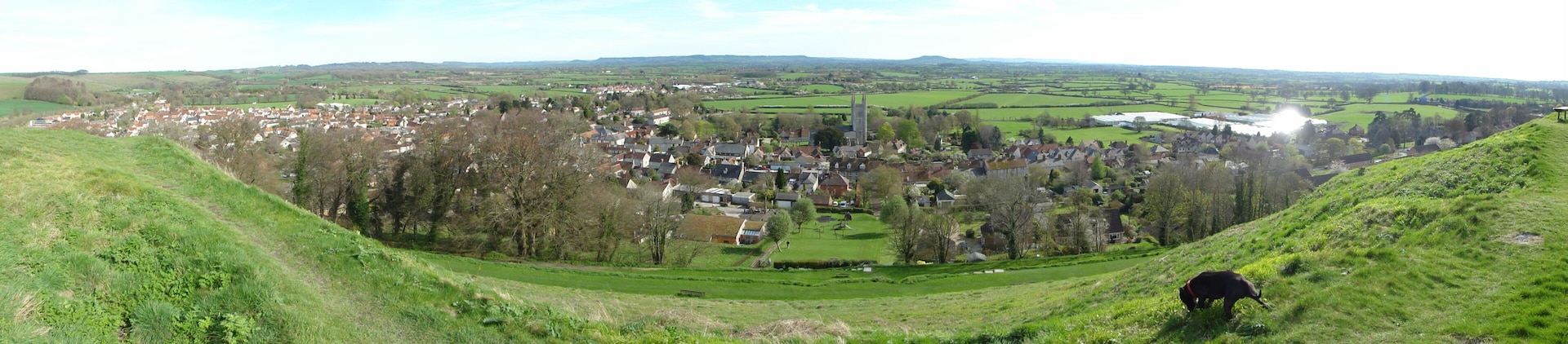 A village nestled in a valley with a prominent church tower, surrounded by green fields and hills.
