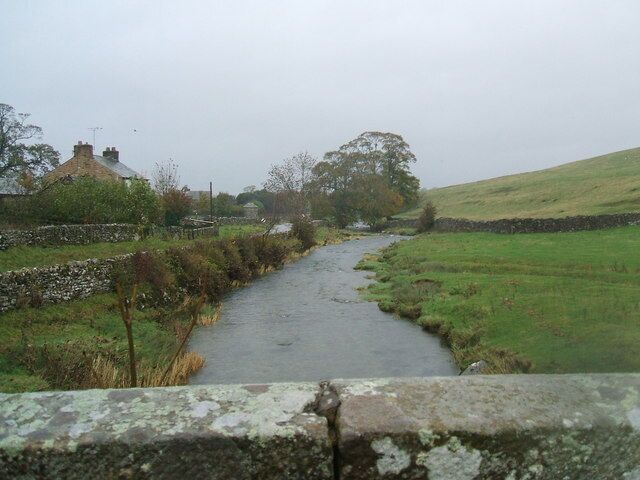 A narrow rural road flanked by stone walls and grassy fields, leading to a house with a chimney.