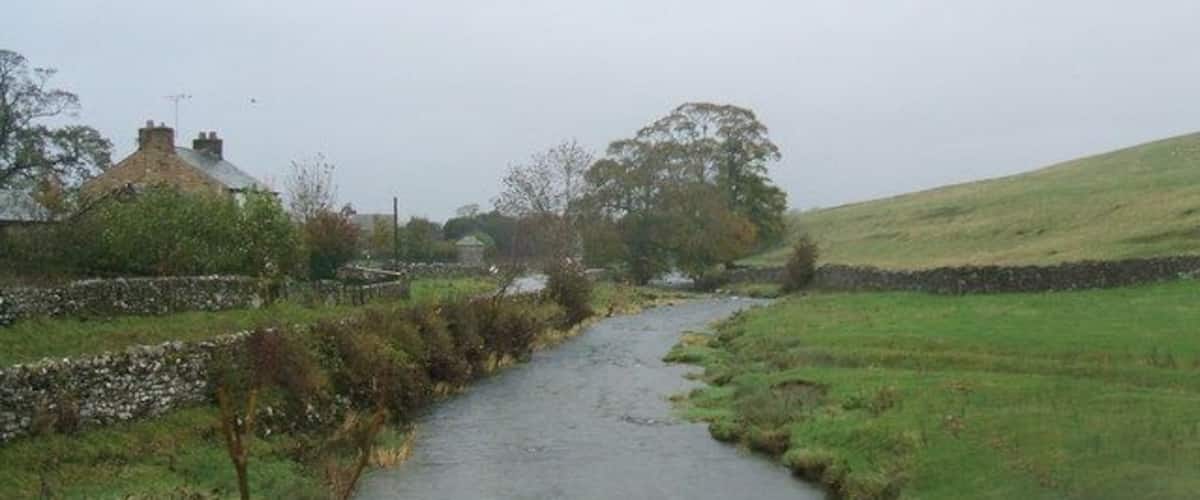 A narrow rural road flanked by stone walls and grassy fields, leading to a house with a chimney.