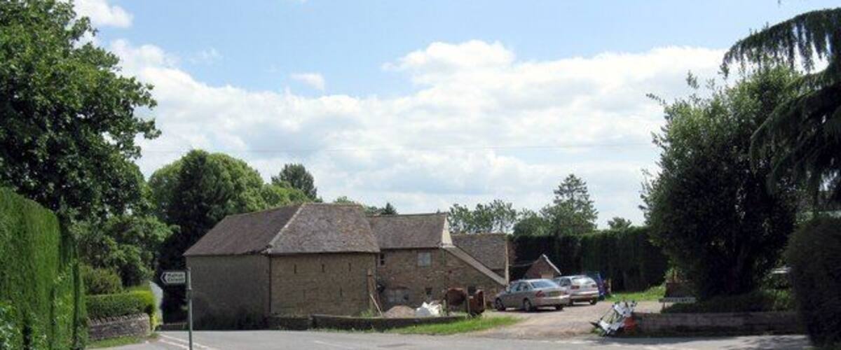 A rural road with a stone building, parked cars, and a grassy area.