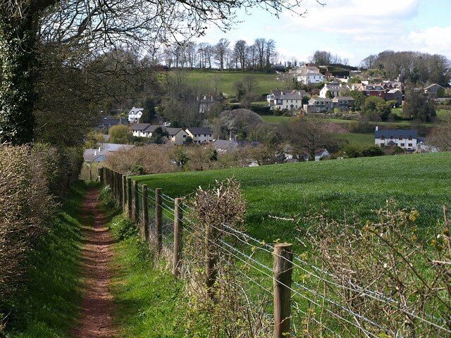 A rural pathway leading through a green field, with houses in the background on a hillside.