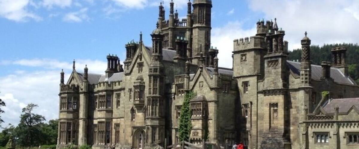 A large, historic stone building with multiple towers and turrets, surrounded by a well-maintained lawn and a stone wall.
