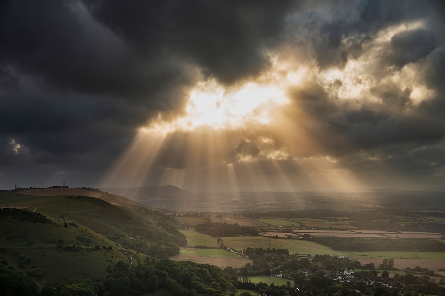 A landscape with rolling hills, a valley, and a distant mountain range under a cloudy sky with sunlight breaking through.