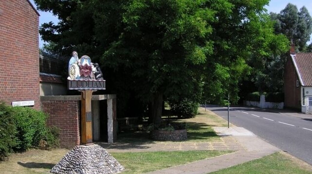 Village sign, Ludham. Just off the main crossroads - which the street sign says is Baker's Arms Green, although the pub is now the King's Arms. The plaques read "In 1019 King Canute gave the Manor of Ludham to the Abbey of St Benet at Holme. St Catherine, whose emblem is a wheel, is the patron saint of the parish church. Design by Joan M Snelling. Carving by Philip Freeman Sayer, 1970"; "Moulded in 1990 by Michael E Fuller from the original". Related geographs: 126957 and 44977.