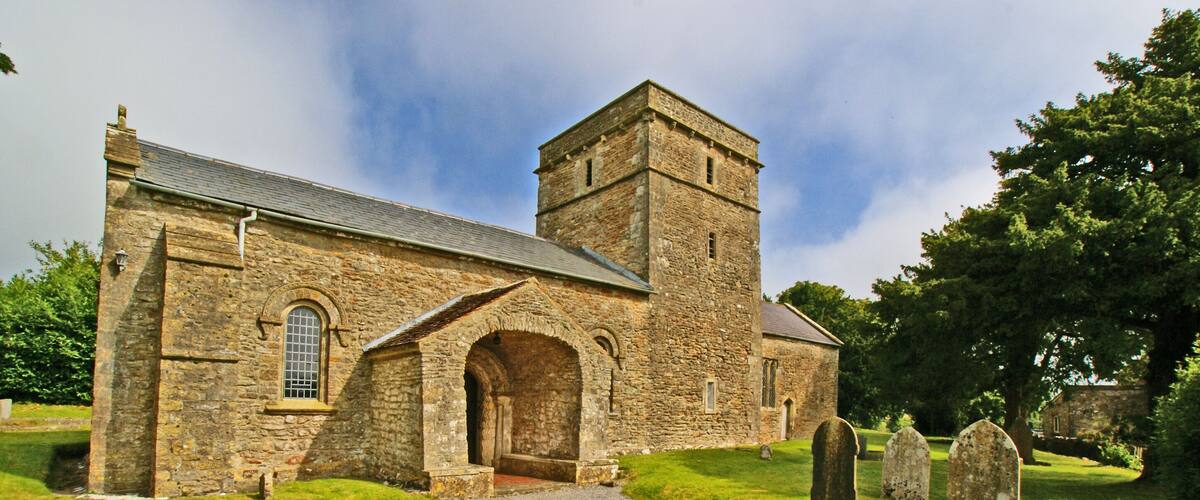 A stone church with a tower and arched entrance, surrounded by a cemetery with headstones.