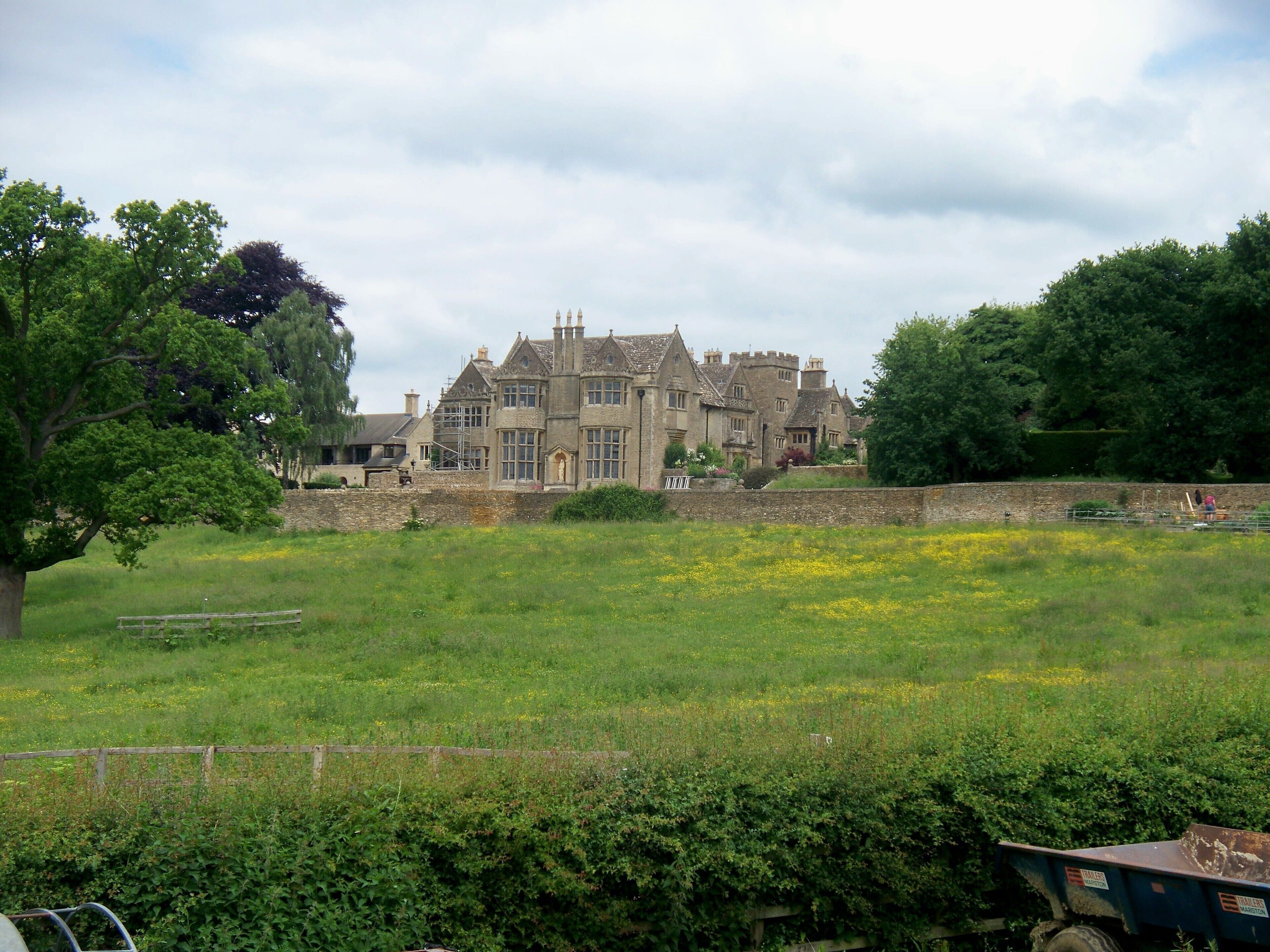 A large stone manor with a well-maintained garden and a field of yellow flowers in the foreground.