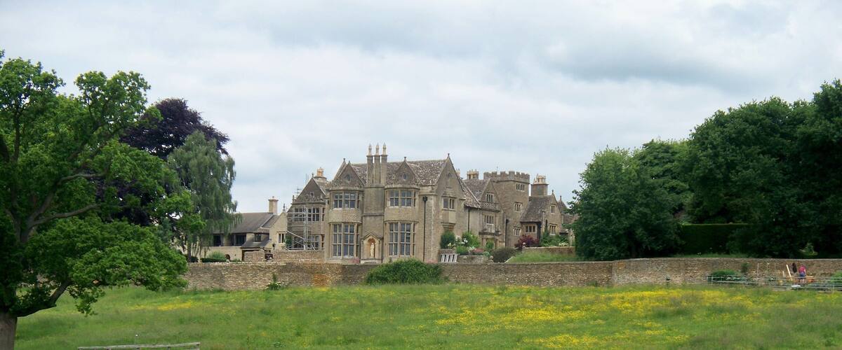 A large stone manor with a well-maintained garden and a field of yellow flowers in the foreground.