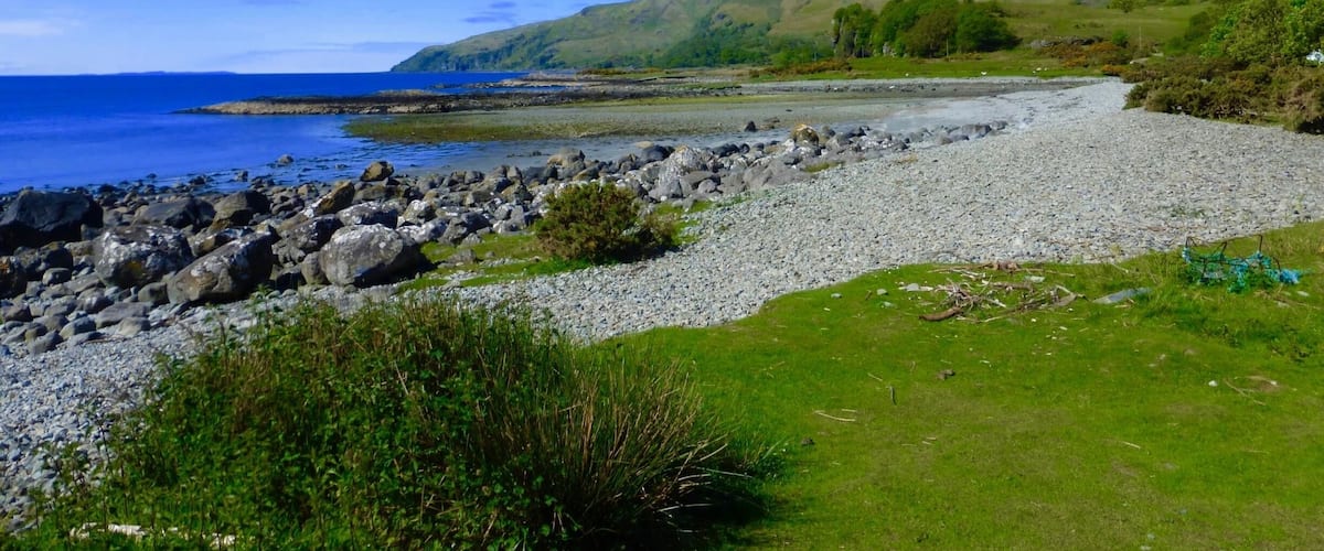 A rocky shoreline with a grassy foreground, a clear blue sky, and distant mountains.