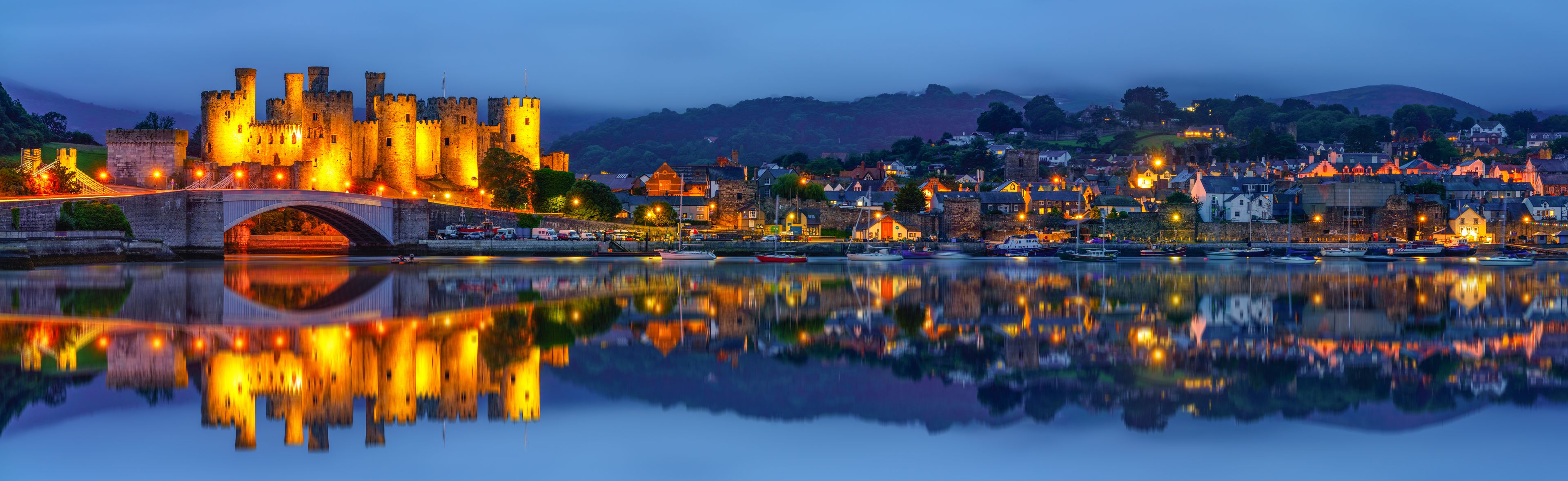 A historic castle with a bridge, a town with buildings, and a calm body of water reflecting the scene.