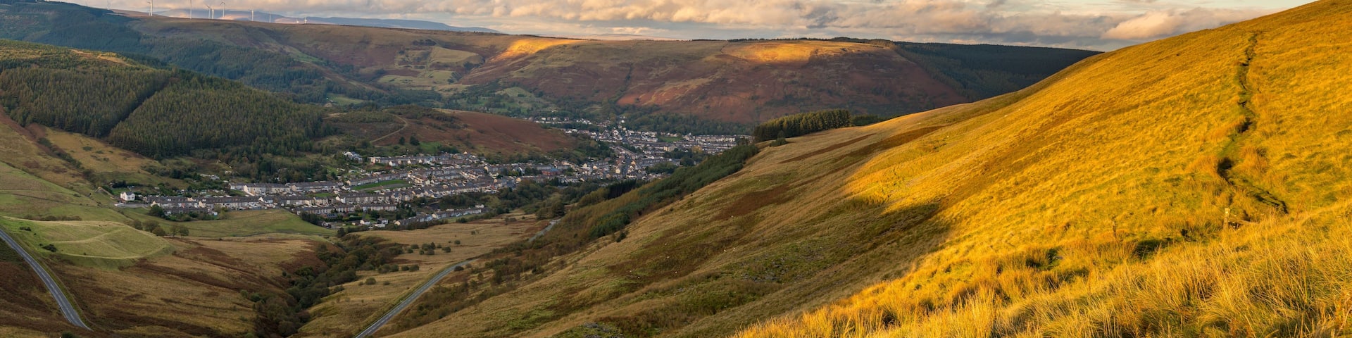A valley with a town nestled among rolling hills, a winding road, and wind turbines in the distance.