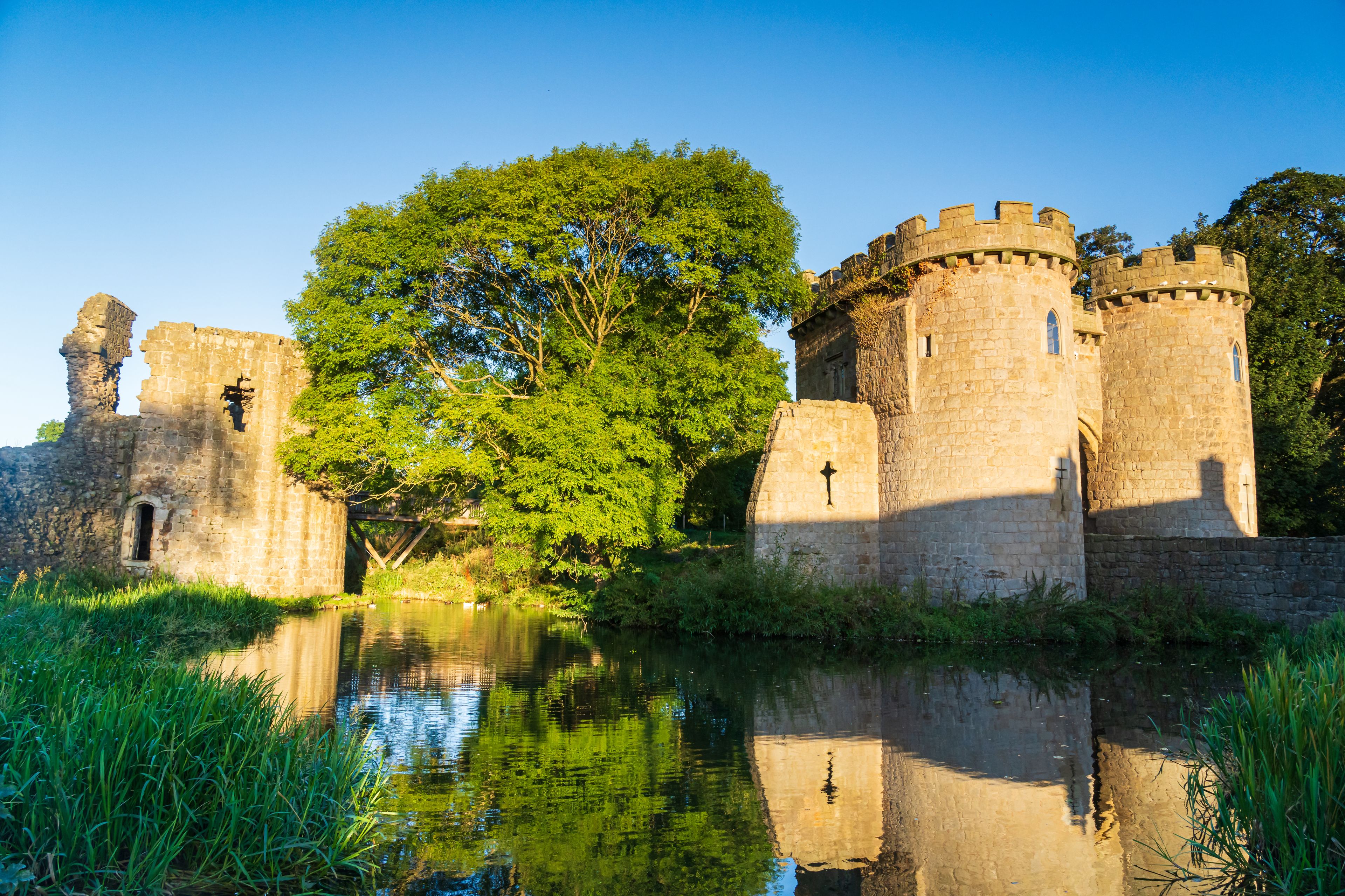 Un castillo histórico con un foso, rodeado de exuberante vegetación y un cielo azul despejado.
