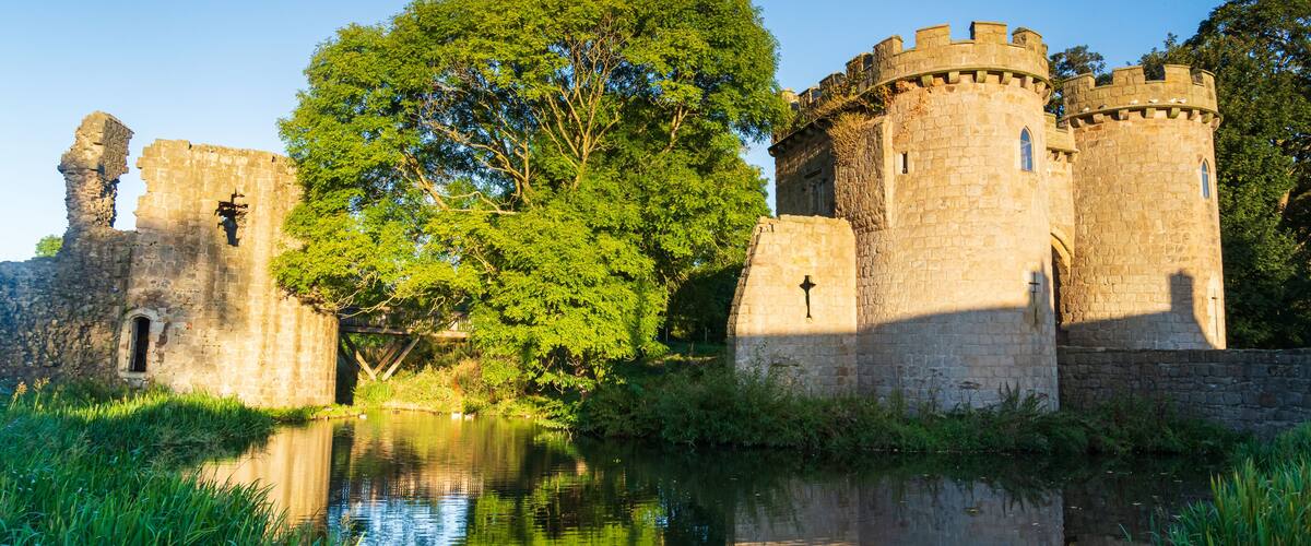 Un castillo histórico con un foso, rodeado de exuberante vegetación y un cielo azul despejado.