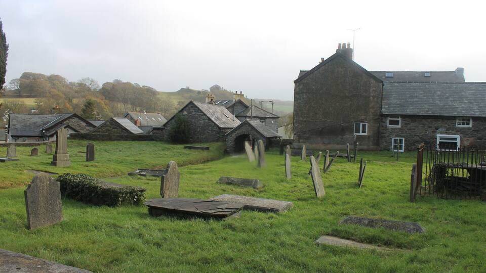 A rural cemetery with stone headstones, a stone building, and a grassy field.
