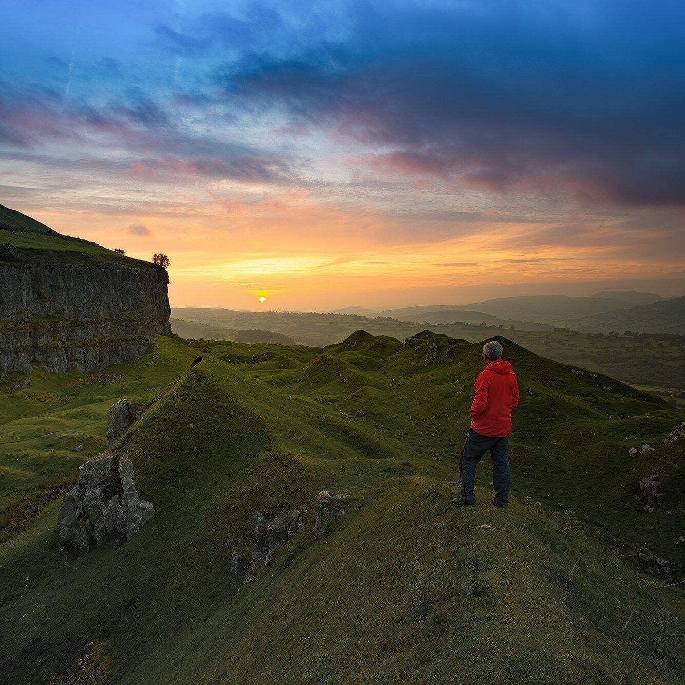 A person in a red jacket standing on a grassy hill overlooking a cliff and a sunset.