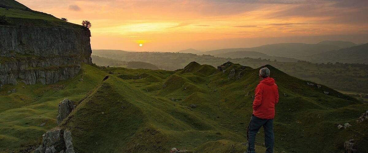 A person in a red jacket standing on a grassy hill overlooking a cliff and a sunset.