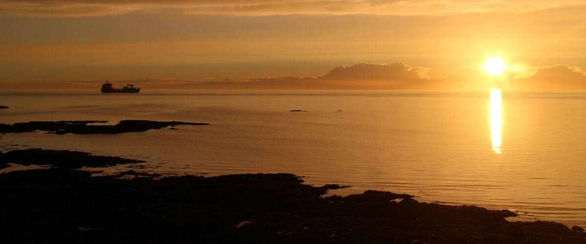 A sunset over a calm sea with a ship in the distance and rocky shoreline.