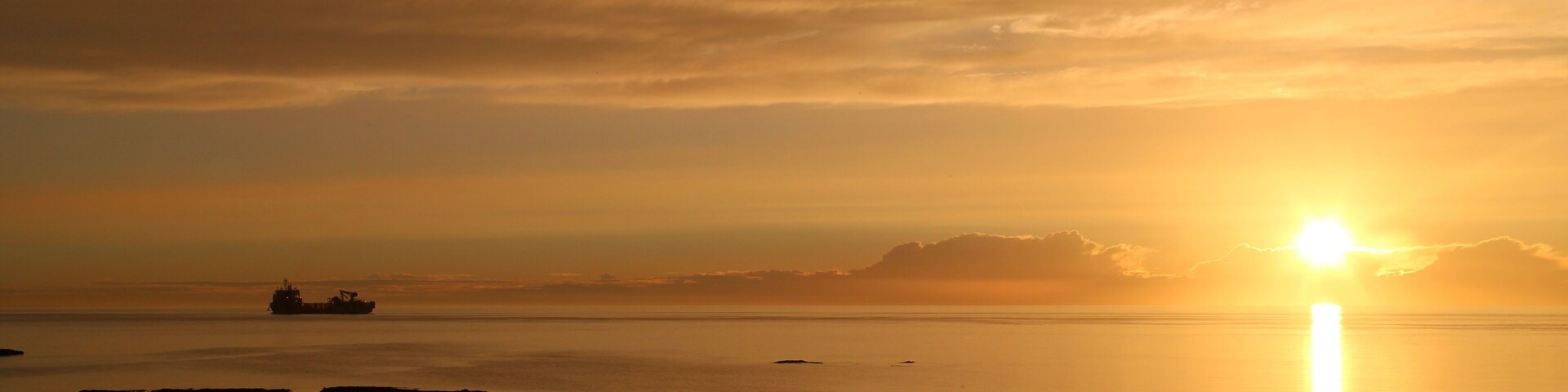 A sunset over a calm sea with a ship in the distance and rocky shoreline.