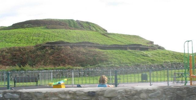 A green hill with a fortification on top, a playground with equipment, and a stone wall in the foreground.