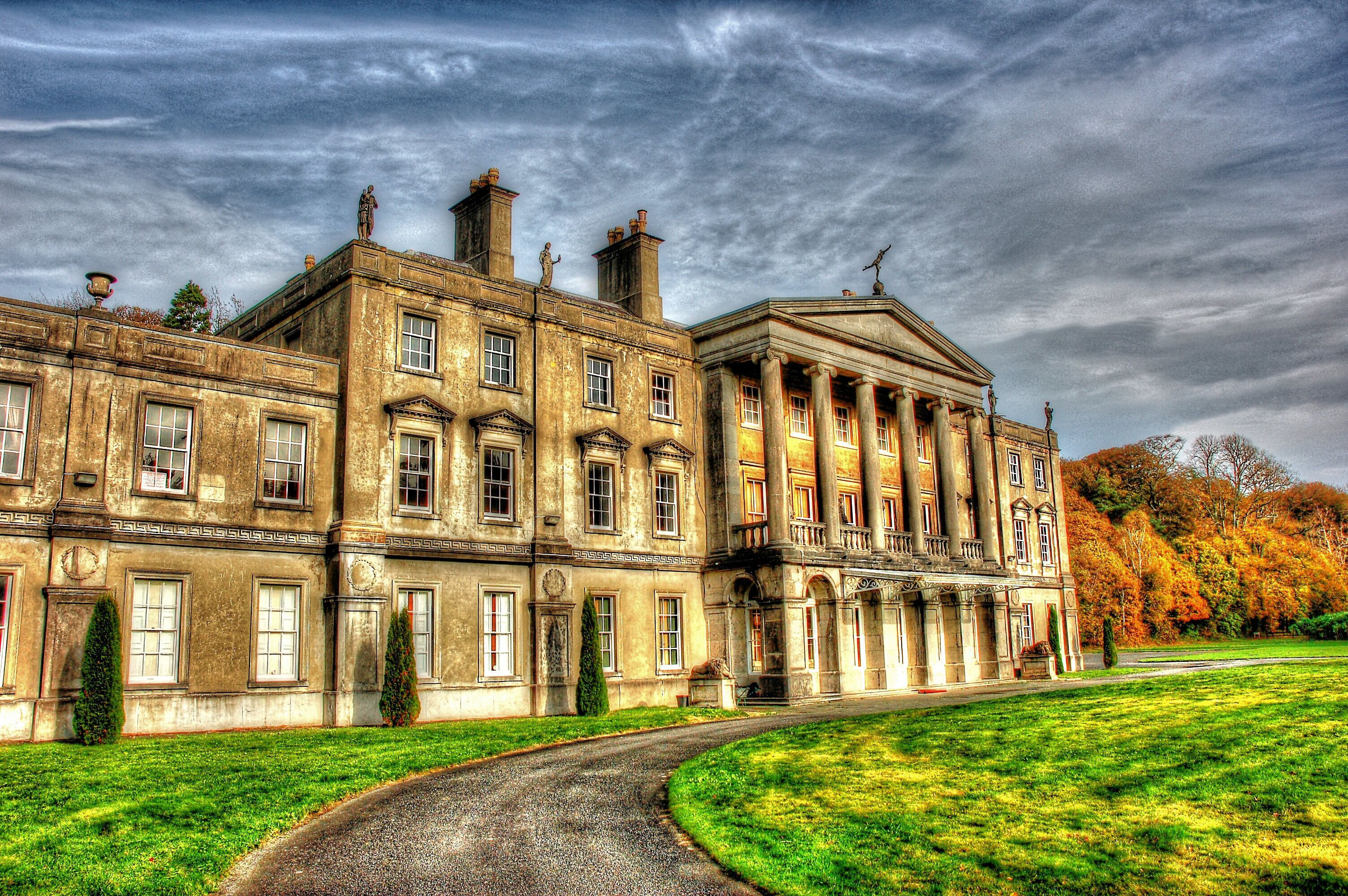A large historic building with classical architecture, surrounded by a well-maintained lawn and trees with autumn foliage.