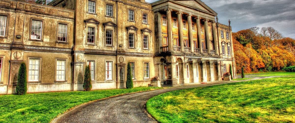 A large historic building with classical architecture, surrounded by a well-maintained lawn and trees with autumn foliage.