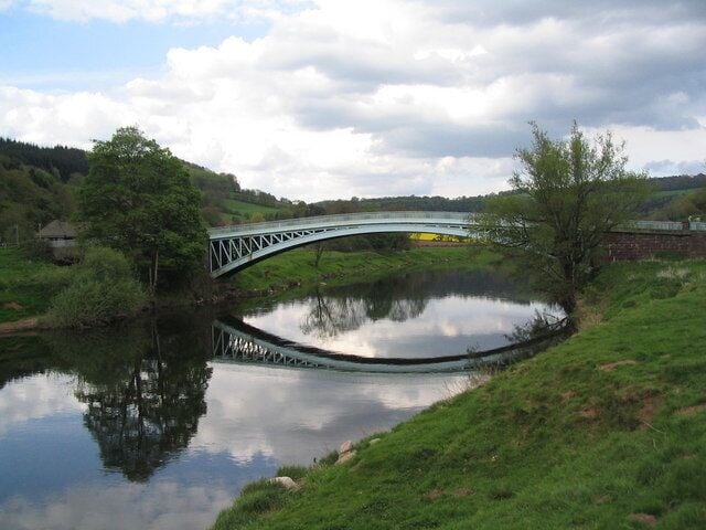 Un puente metálico sobre un río tranquilo, rodeado de colinas y árboles verdes.