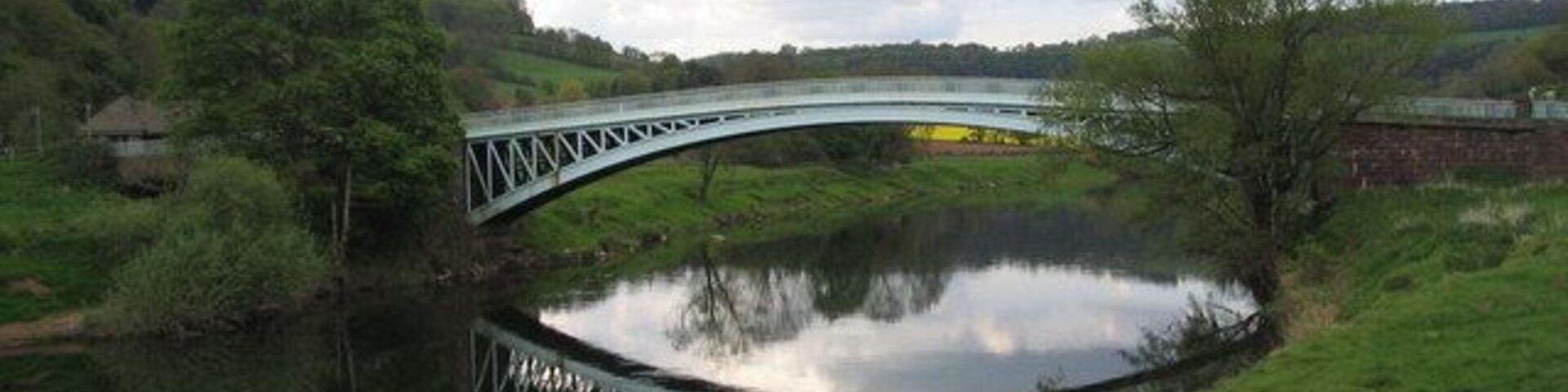 Un puente metálico sobre un río tranquilo, rodeado de colinas y árboles verdes.