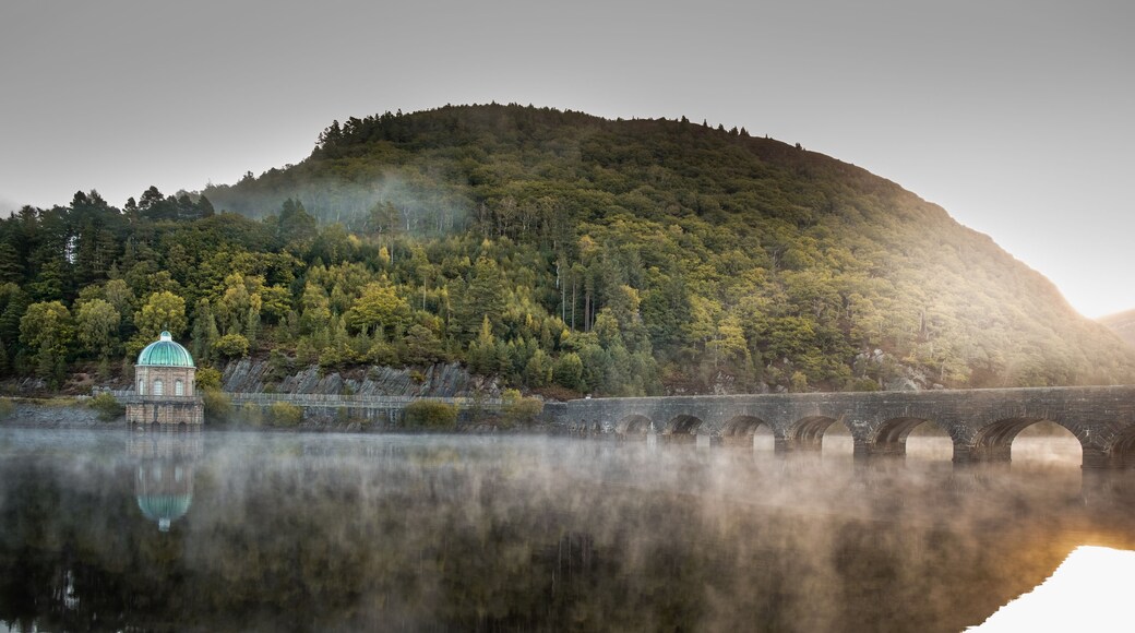 A serene lake with a stone bridge, a small building with a dome, and a forested hill in the background.