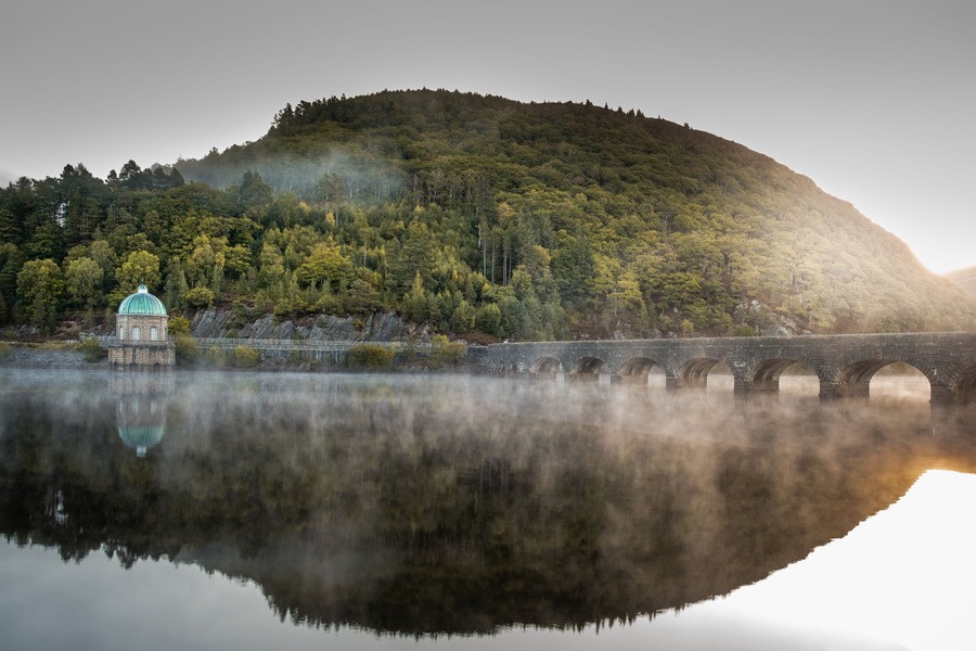 A serene lake with a stone bridge, a small building with a dome, and a forested hill in the background.
