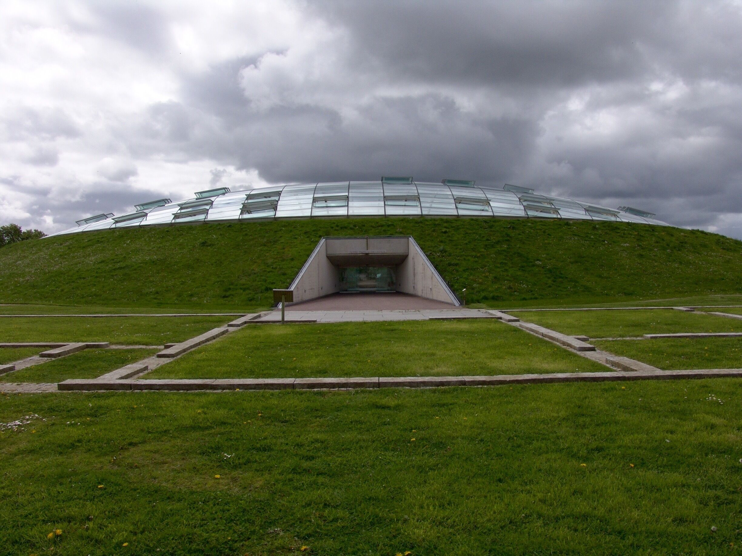 A modern building with a glass facade on a grassy hill under a cloudy sky.