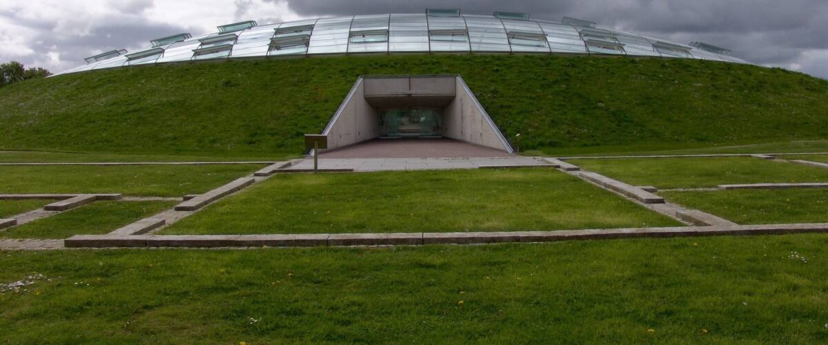 A modern building with a glass facade on a grassy hill under a cloudy sky.