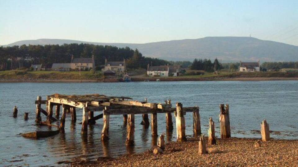 A wooden pier extending into calm water, surrounded by a pebbled shore and a backdrop of hills and houses.