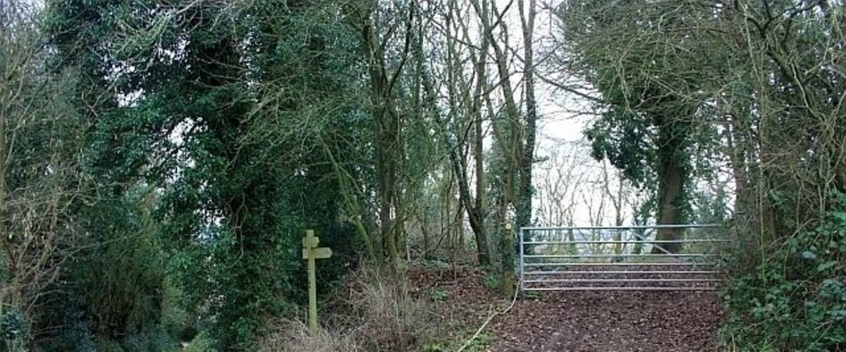 A narrow, unpaved path leading through a wooded area with a metal gate and a directional signpost.