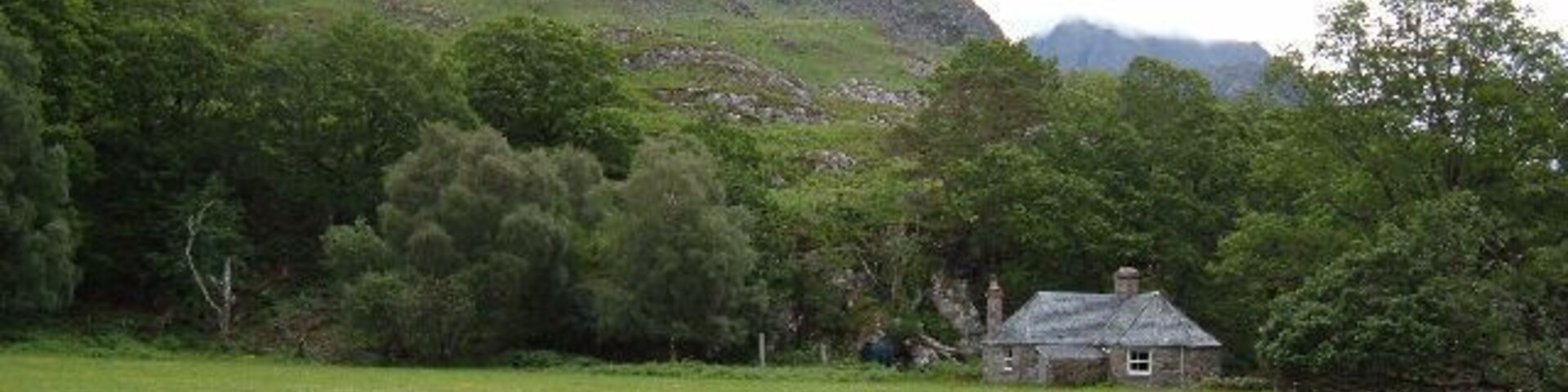 Paisagem rural com um campo gramado, uma casa de pedra e uma cordilheira ao fundo.