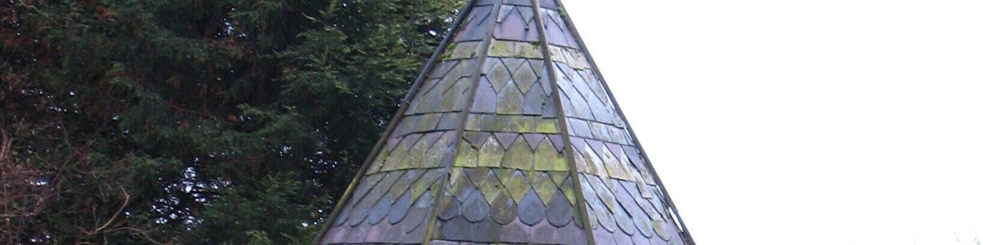 A stone gazebo with a pointed roof and a weather vane atop.