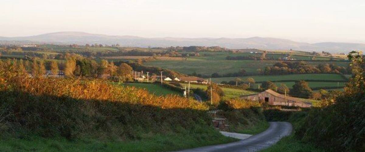 A rural road winding through green fields with distant hills and a few buildings.
