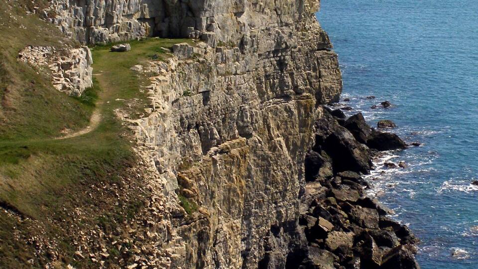 A coastal cliff with a path leading to a grassy area and a rocky shoreline.