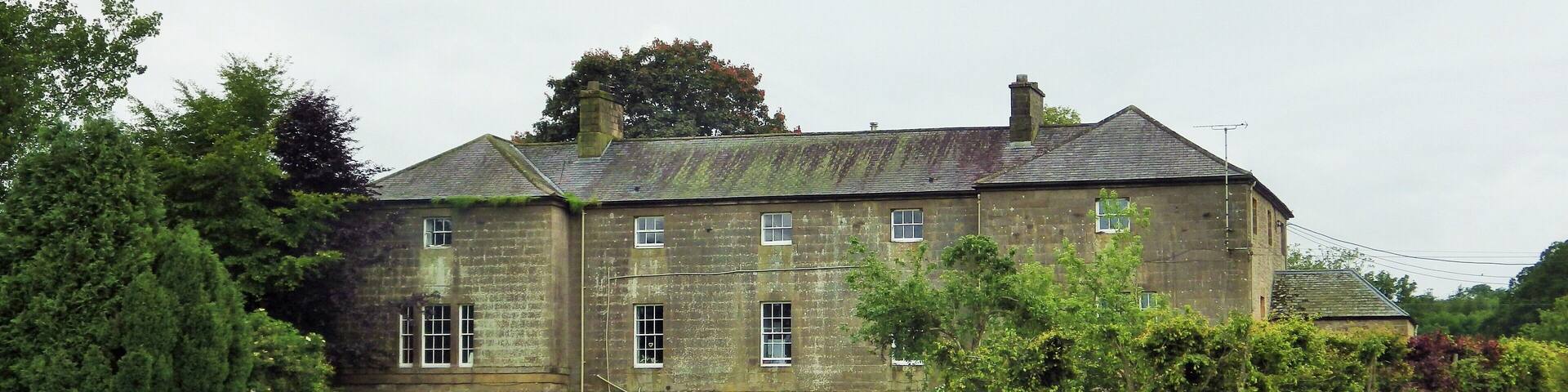 A large stone house with multiple windows, surrounded by lush greenery and a grassy field.