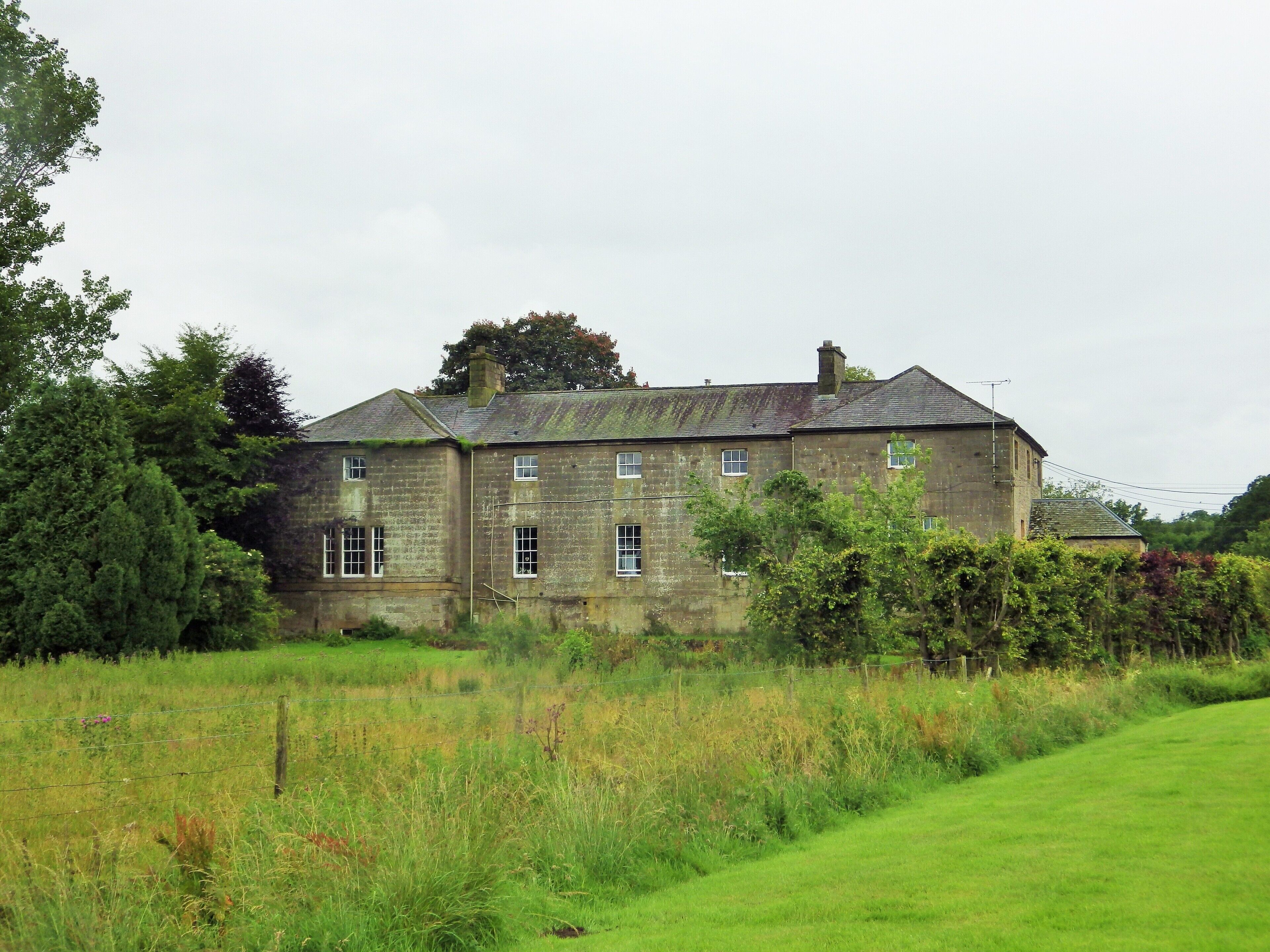 A large stone house with multiple windows, surrounded by lush greenery and a grassy field.