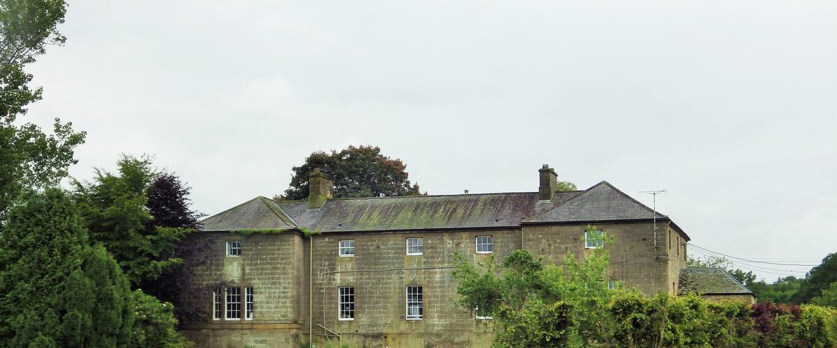 A large stone house with multiple windows, surrounded by lush greenery and a grassy field.