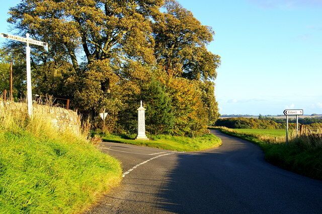 A rural road with a white signpost, a stone wall, and a large tree.