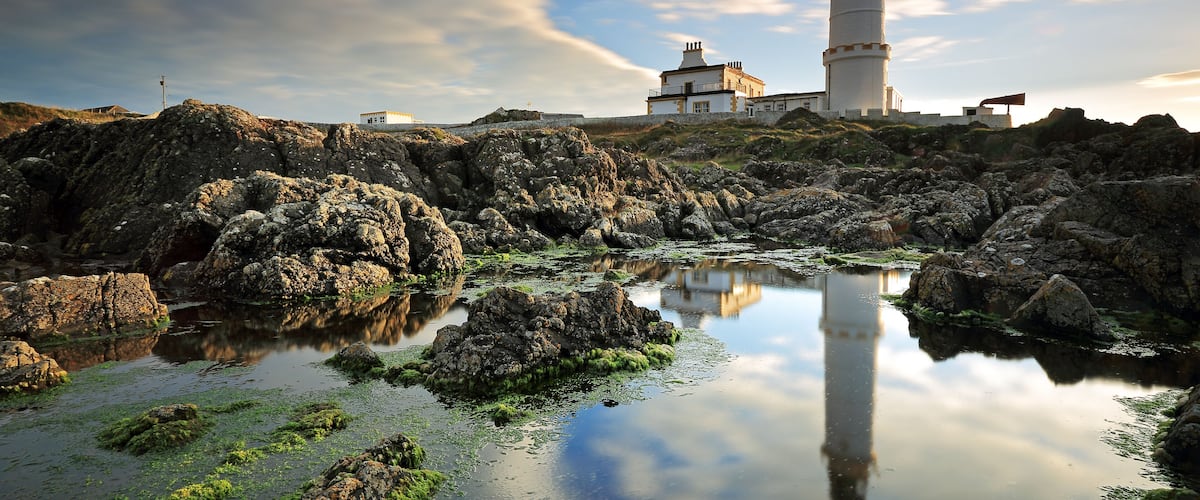 A coastal landscape with a lighthouse, rocky terrain, and a calm reflective water body.