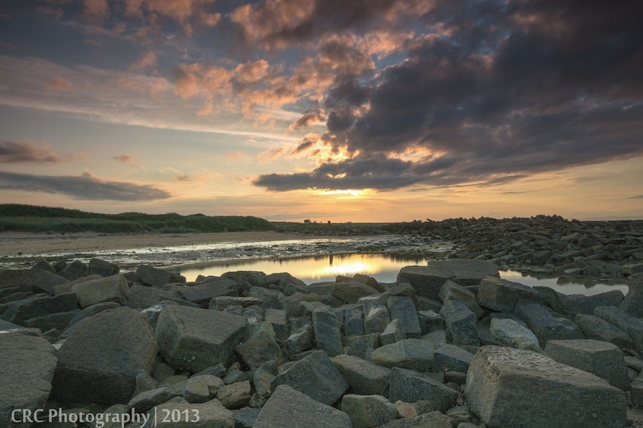 A rocky shoreline with a calm body of water reflecting the sunset sky.