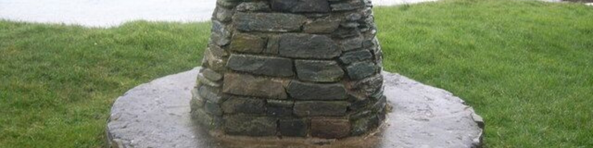 Monumento de piedra con una placa, rodeado de hierba y un cuerpo de agua.
