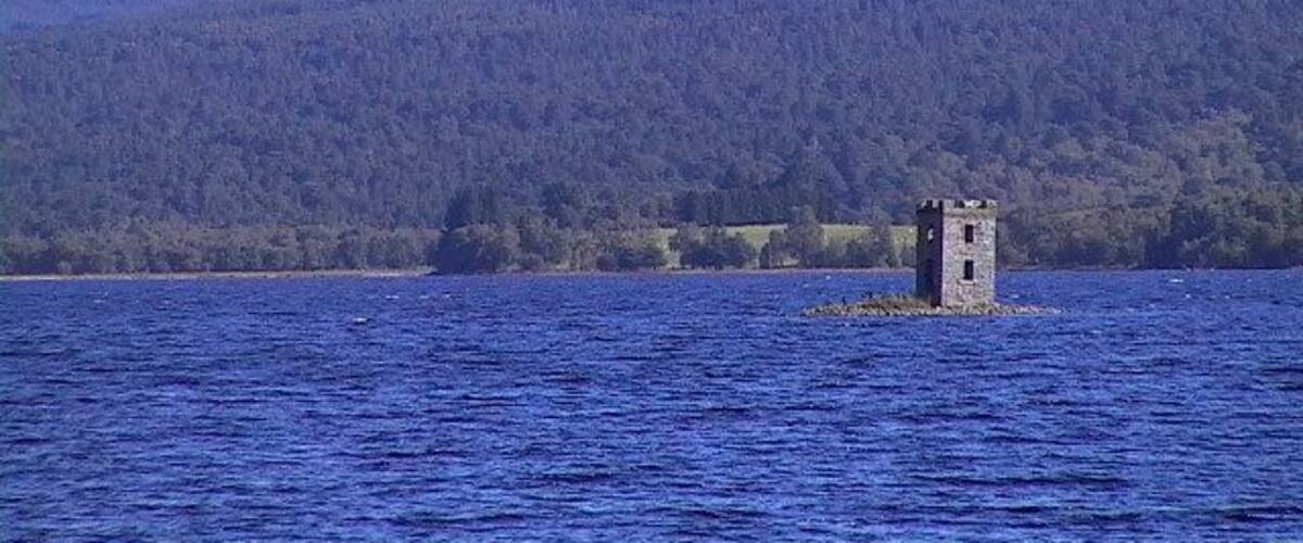 Een toren op een klein eiland in een meer, met bergen op de achtergrond.
