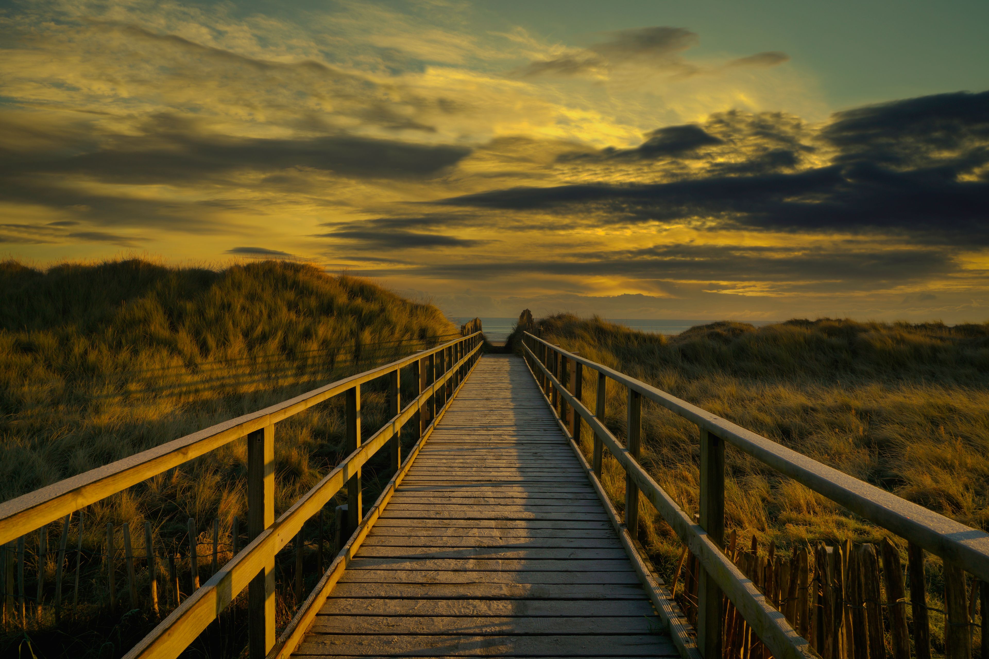 A wooden boardwalk leading through a grassy landscape with a cloudy sky.
